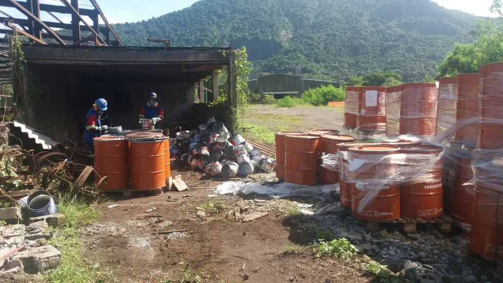 Funcionários com EPI manuseando tambores de resíduos perigosos em área contaminada