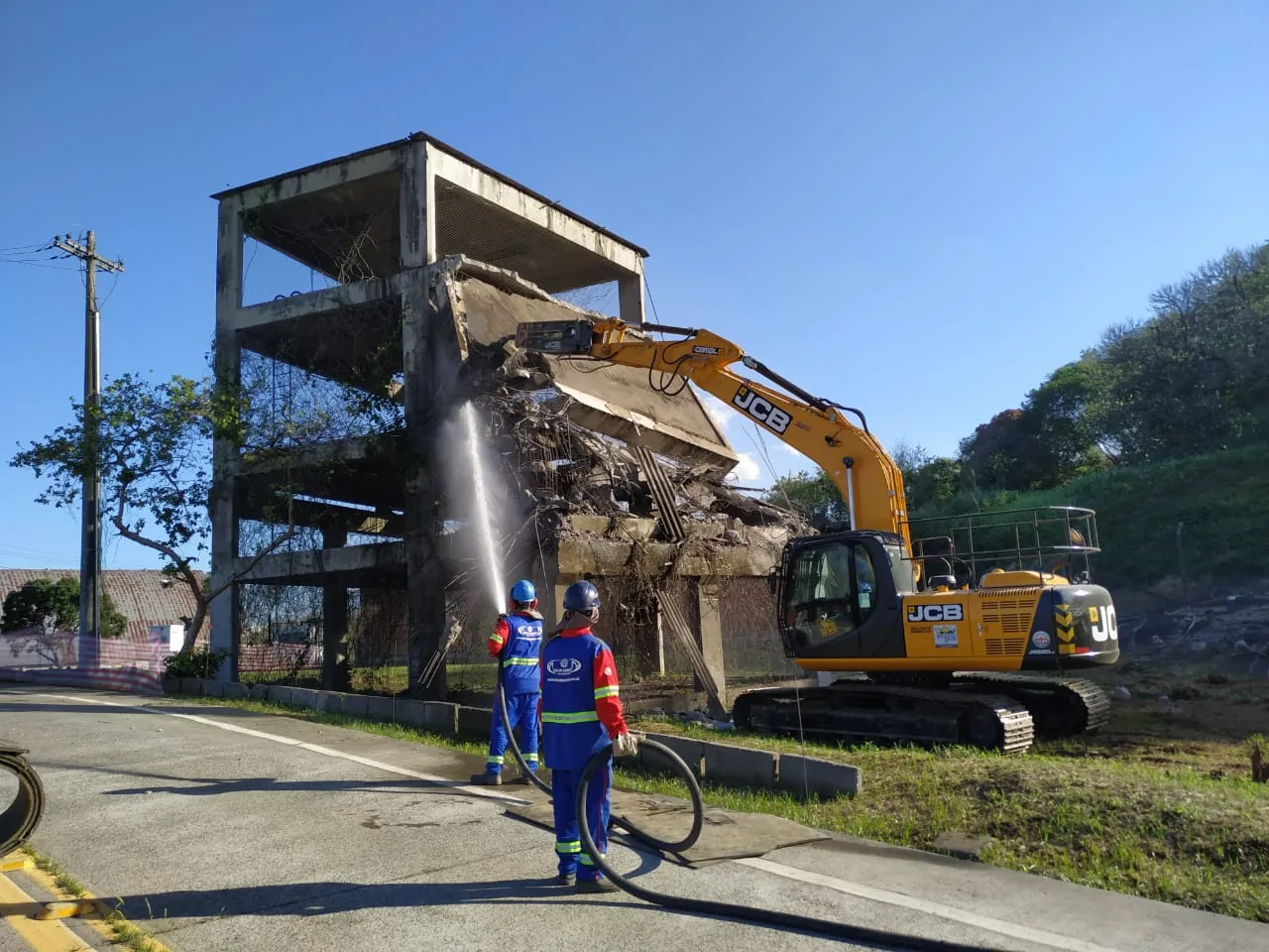 Escavadeira JCB em demolição mecânica de edificação com equipe fazendo aspersão de água